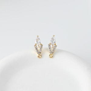 May include: A pair of gold-colored earrings with a V-shaped design. Each earring features a teardrop-shaped clear gemstone at the top, with smaller clear stones set along the V. A small loop is at the bottom of each earring.