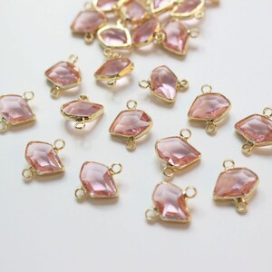 K&ouml;nnte beinhalten: Eine Sammlung von rosafarbenen, facettierten Glas-Verbindungsanh&auml;ngern mit goldfarbenen Metallscharnieren. Die Anh&auml;nger sind diamantf&ouml;rmig und haben ein facettierten Design.
