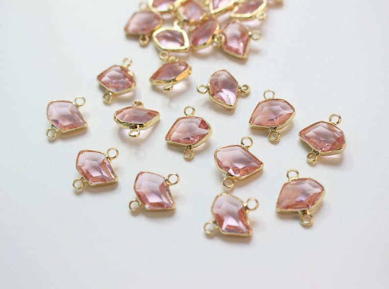 K&ouml;nnte beinhalten: Eine Sammlung von rosafarbenen, facettierten Glas-Verbindungsanh&auml;ngern mit goldfarbenen Metallscharnieren. Die Anh&auml;nger sind diamantf&ouml;rmig und haben ein facettierten Design.