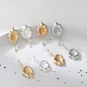 Puede incluir: Primer plano de varios hallazgos de joyería en forma de hoja, en tonos dorados y plateados. Cada hoja tiene un patrón de vetas detallado y un bucle para sujetar a otros componentes de joyería. Los hallazgos están dispuestos sobre una superficie blanca.