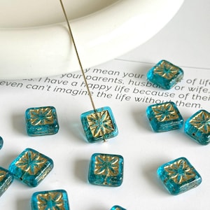 Op de afbeelding: Vierkante turquoise glazen kralen met een gouden bloemenpatroon. De kralen zijn geregen op een dunne draad en liggen verspreid op een wit oppervlak. De kralen zijn ongeveer 1,3 cm vierkant.