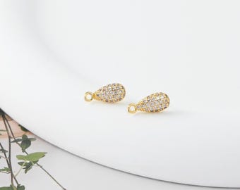 May include: Two gold-colored teardrop-shaped jewelry charms, encrusted with small, clear crystals. Each charm has a small loop at the top for attachment. The charms are displayed on a white, curved surface.