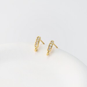 May include: A pair of gold-colored stud earrings. Each earring features a rectangular design with a row of small, clear gemstones. The earrings have a post and a small loop at the bottom.