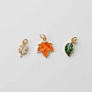 May include: Three gold-colored charms with different designs. One is a four-leaf clover with clear stones, another is an orange maple leaf, and the last is a green leaf. Each charm has a small loop for attaching to a necklace or bracelet.