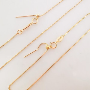 Puede incluir: Dos cadenas de oro con longitudes ajustables. Cada cadena tiene una pequeña bola de oro en el extremo y un cierre. Las cadenas están sobre un fondo blanco.