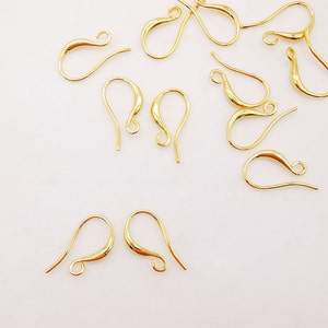 Puede incluir: Un grupo de ganchos para pendientes de color dorado, también conocidos como alambres para pendientes, están dispersos sobre una superficie blanca. Los ganchos son de metal y tienen forma de lágrima.