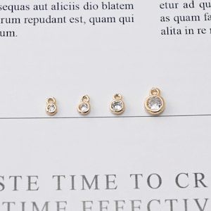 May include: Four gold-colored charms with a small, clear, round gemstone in the center. The charms are in a row, each slightly larger than the last. The charms have a loop at the top for attaching to jewelry.