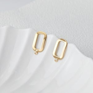 May include: Two gold-coloured rectangular earring charms with a small loop at the bottom for attaching to earring hooks. The charms are displayed on a white, scalloped surface.