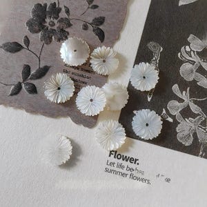 Könnte beinhalten: Eine Ansammlung von acht runden weißen Knöpfen mit einem geschnitzten Blütenblattmuster und einem Mittelloch. Die Knöpfe sind auf einer Oberfläche angeordnet, die den gedruckten Text Flower. Let life be bea summer flowers. aufweist.