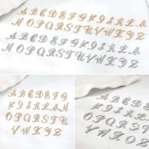 Pode incluir: Uma coleção de pingentes de letras do alfabeto em metal dourado e prateado com uma fonte cursiva. Cada letra apresenta uma superfície texturizada com pequenas pedras transparentes incrustadas. Os pingentes estão dispostos em conjuntos completos de A a Z.