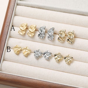 May include: Two pairs of gold earrings with a textured, abstract design. The earrings are displayed in a wooden box with a white fabric lining. The box is labeled with the letters A and B.
