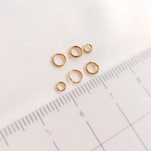 May include: Five gold-colored metal jump rings of varying sizes arranged near a ruler. The ruler shows the rings range from approximately 0.15 inches to 0.25 inches in diameter. Each ring features a small opening, indicating they are split rings.