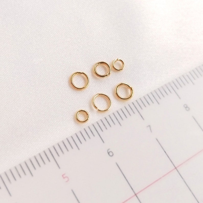 May include: Five gold-colored metal jump rings of varying sizes arranged near a ruler. The ruler shows the rings range from approximately 0.15 inches to 0.25 inches in diameter. Each ring features a small opening, indicating they are split rings.