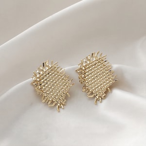 May include: A pair of gold-toned earrings with a woven, textured design. The earrings have a square shape with frayed edges.