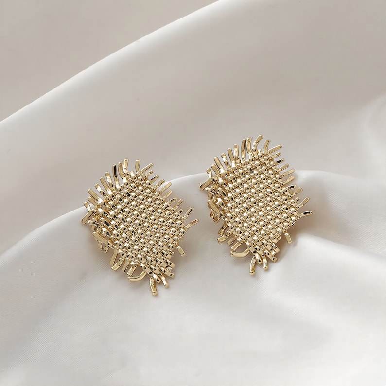 May include: A pair of gold-toned earrings with a woven, textured design. The earrings have a square shape with frayed edges.