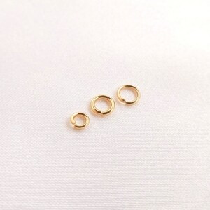 May include: Three gold-colored metal jump rings of varying sizes arranged in a diagonal line. Each circular ring features a small split or opening, characteristic of jewelry findings used for connecting components in various craft projects.