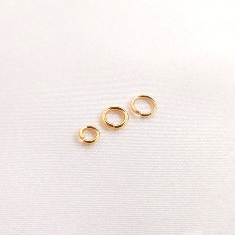 May include: Three gold-colored metal jump rings of varying sizes arranged in a diagonal line. Each circular ring features a small split or opening, characteristic of jewelry findings used for connecting components in various craft projects.