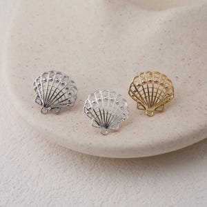 Peut inclure: Trois breloques en forme de coquillage, argentées et dorées. Les breloques présentent un design ajouré délicat avec une petite boucle en bas pour la fixation. Elles sont présentées sur une surface de couleur neutre.