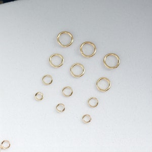 May include: A collection of gold jump rings of various sizes, arranged on a white surface.