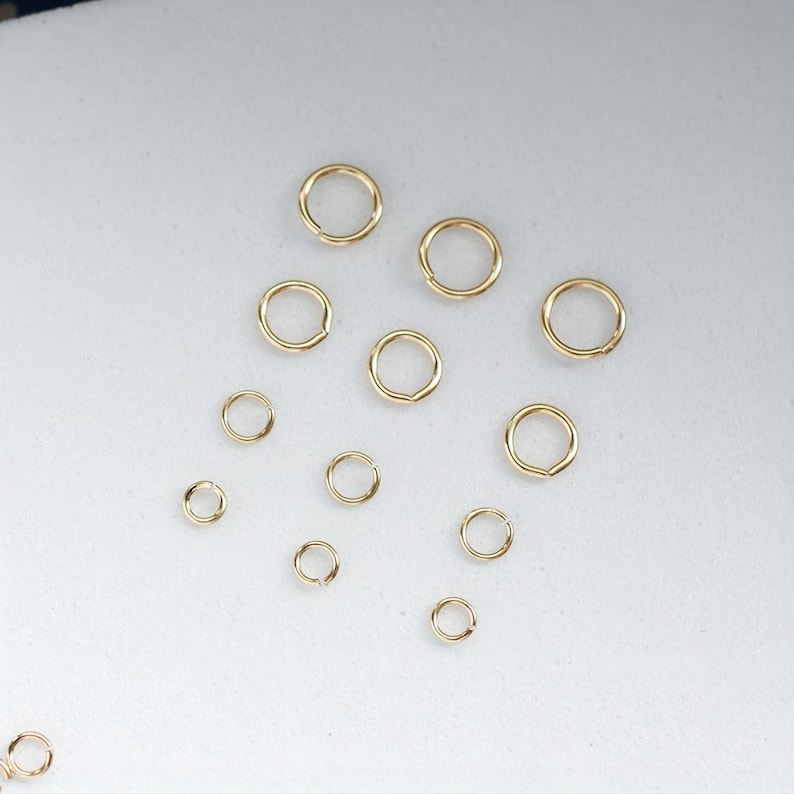May include: A collection of gold jump rings of various sizes, arranged on a white surface.