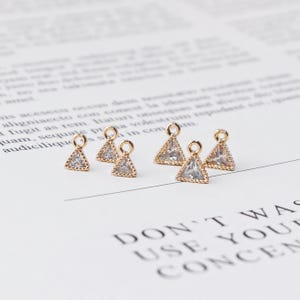 Könnte beinhalten: Sechs goldfarbene Dreiecksanhänger mit klaren, funkelnden Steinen. Jeder Anhänger hat oben eine kleine Schlaufe zum Befestigen an Schmuck. Die Anhänger sind auf einer weißen Oberfläche mit schwachem Text im Hintergrund angeordnet.