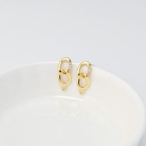 Puede incluir: Dos colgantes ovalados dorados y enlazados con pequeñas piedras transparentes. Cada colgante tiene un pequeño lazo en la parte inferior. Los colgantes se muestran sobre una superficie blanca.