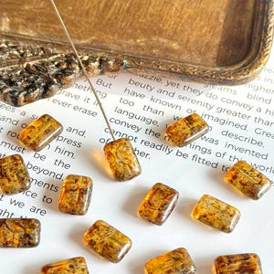 May include: Close-up of several rectangular amber-colored beads with a textured surface. The beads are scattered on a white surface with printed text and a decorative wooden tray in the background. A needle is inserted into one of the beads.