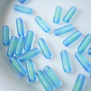 Pode incluir: Uma coleção de contas translúcidas e alongadas em tons de azul e verde. As contas têm uma forma arredondada, semelhante a uma pílula, e parecem ser feitas de vidro ou material semelhante. Estão espalhadas sobre uma superfície branca.