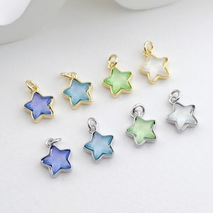 Puede incluir: Colección de colgantes con forma de estrella en varios colores, incluyendo azul, verde y blanco. Los colgantes tienen un borde metálico en oro o plata y un pequeño lazo para sujetar. Las estrellas son el tema.