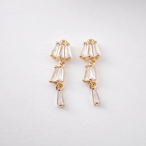 Op de afbeelding: Een paar goudkleurige oorhangers. Elke oorbel heeft een cluster van heldere, rechthoekig geslepen kristallen aan de bovenkant, met twee extra kristalclusters en een enkele kristallen hanger die eronder hangt. Op een witte achtergrond.
