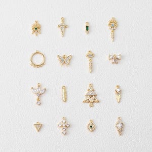 May include: A collection of gold-toned jewelry charms, including crosses, butterflies, and geometric shapes. Some charms are adorned with clear and green gemstones. The charms are designed for jewelry making and crafting.