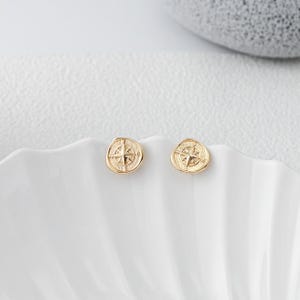 Puede incluir: Dos pendientes dorados. Cada pendiente presenta un diseño de rosa de los vientos. Los pendientes son redondos con un borde ligeramente irregular. Se muestran sobre una superficie blanca y festoneada.