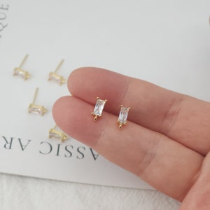 May include: Gold-colored stud earrings with rectangular, clear crystal stones. The earrings are displayed on a light background, with several pairs visible. The studs have a simple, elegant design.