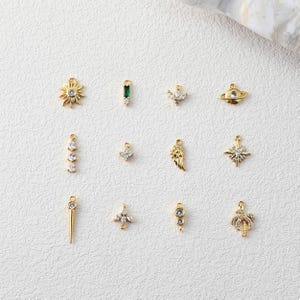 Puede incluir: Una colección de colgantes dorados con varios diseños, incluyendo un sol, un ala, una estrella y una palmera. Cada colgante está adornado con piedras preciosas claras y verdes. Los colgantes se muestran sobre una superficie blanca.