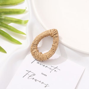 May include: A teardrop-shaped earring made of woven, light brown material. The earring is on a white surface with a card that reads "Favorite Flowers". The earring is a natural, bohemian style.