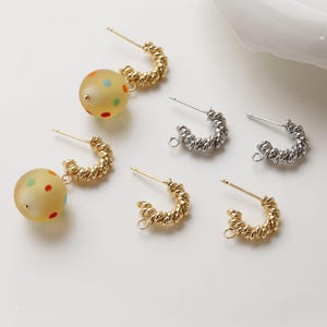 May include: Earrings in gold and silver. Some have a textured hoop design, others feature a yellow ball with colourful polka dots. The earrings are displayed on a white surface. Measurements are in centimetres.