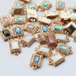 May include: A collection of gold-colored rectangular charms with iridescent centers in various colors, including white, pink, blue, and green. Each charm is framed with small, clear crystals and has loops for attaching to jewelry.