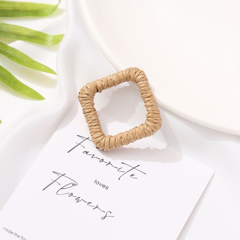 May include: A square-shaped earring with a woven, natural fiber design. The earring is a light tan color and sits on a white card that reads "Favorite Flowers". The background is a white fabric.