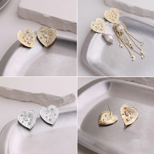 May include: A collection of heart-shaped earrings featuring textured gold and silver finishes with embedded clear crystals. One pair includes dangling gold chains adorned with small star and moon charms and a white teardrop pearl accent.