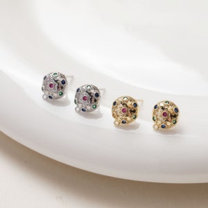 May include: Three pairs of gold and silver stud earrings with a round design featuring a red, green, and blue gemstone pattern.