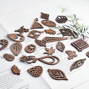 Op de afbeelding: Een verzameling houten uitsparingen in verschillende vormen, waaronder bladeren, druppels en geometrische ontwerpen. De uitsparingen zijn warmbruin van kleur en liggen op een wit oppervlak, wat wijst op een knutselproject.