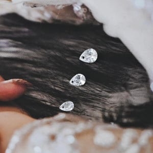 以下が含まれることがあります： 上部に小さな穴がある、透明な涙型のラインストーンが3つ。 ラインストーンは、ぼやけた明るい色のパターンを持つ暗い背景に表示されています。 宝石は輝き、光を反射しています。