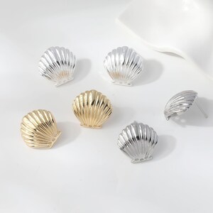 Könnte beinhalten: Eine Sammlung von fünf muschelförmigen Ohrstecksteckern, bestehend aus drei silberfarbenen und zwei goldfarbenen Stücken. Jeder Ohrring hat eine gerippte, fächerartige Textur, die an eine Jakobsmuschel erinnert.