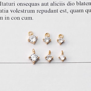 May include: Six small gold-colored charms with clear, sparkling cubic zirconia stones. The charms are in various shapes, including round and square, and are displayed on a white surface. Each charm has a small loop for attaching to jewelry.