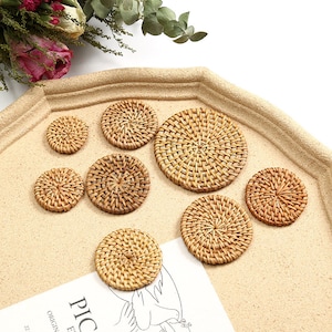 May include: A selection of woven rattan coasters in different sizes. The coasters are circular, with a natural, light brown hue and a woven design. They are displayed on a light tan surface, suitable for home decor.