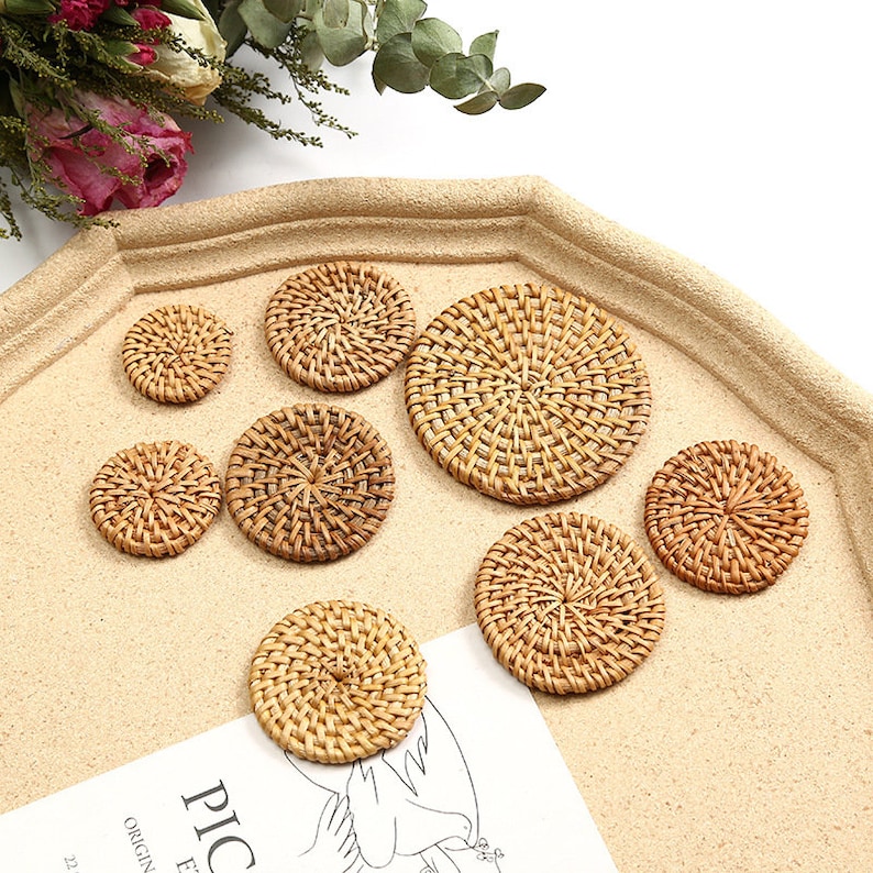 May include: A selection of woven rattan coasters in different sizes. The coasters are circular, with a natural, light brown hue and a woven design. They are displayed on a light tan surface, suitable for home decor.