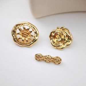 May include: Three gold-toned metal embellishments with floral designs. One is a round, openwork design with a stylized flower in the center. The other two are solid rose designs.
