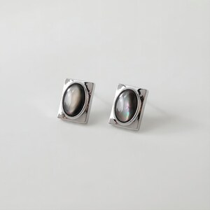 May include: A pair of silver earrings with a square setting and a black oval-shaped stone.