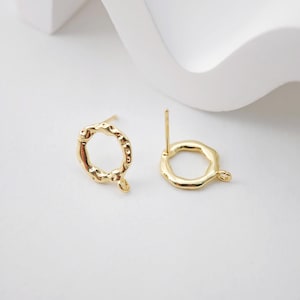 Op de afbeelding: Gouden oorbel-bevestigingen met een getextureerde, onregelmatige cirkelvorm. De bevestigingen hebben een pen en een lus om een ​​bedel of hanger aan te bevestigen.