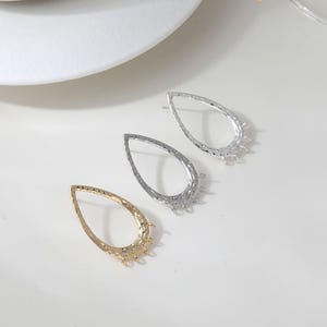 Puede incluir: Tres componentes de pendientes en forma de lágrima en oro, plata y un tercer metal plateado. Cada uno tiene una superficie texturizada y múltiples bucles pequeños en la parte inferior para sujetar cuentas u otros adornos. Los componentes están dispuestos sobre una superficie blanca.